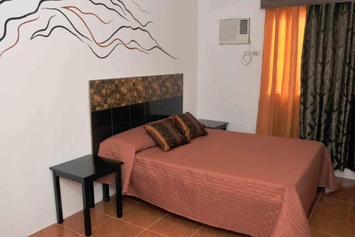 A bedroom with a bed, a nightstand, a wall art piece, and a window with curtains.