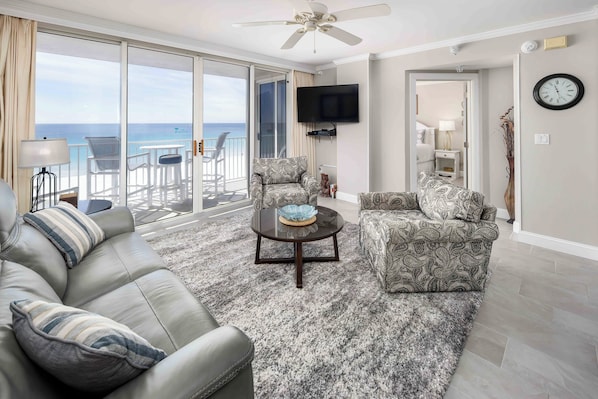 A living room with a sofa, armchair, and coffee table, a TV mounted on the wall, and a view of the ocean through large sliding doors.