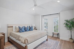 Un dormitorio con cama, ventilador de techo, ventana con persianas, una mesita y una planta en maceta.