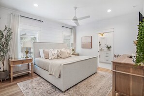 Un dormitorio con cama, mesitas de noche, mesilla auxiliar, ventilador de techo, ventana con persianas, espejo y cómoda.
