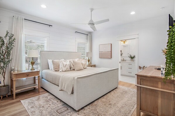 Un dormitorio con cama, mesitas de noche, mesilla auxiliar, ventilador de techo, ventana con persianas, espejo y cómoda.