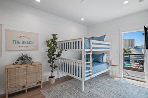 Una cama litera infantil con ropa de cama azul, una mesita de mimbre con una planta, un póster en la pared y vistas de edificios a través de una puerta de cristal.