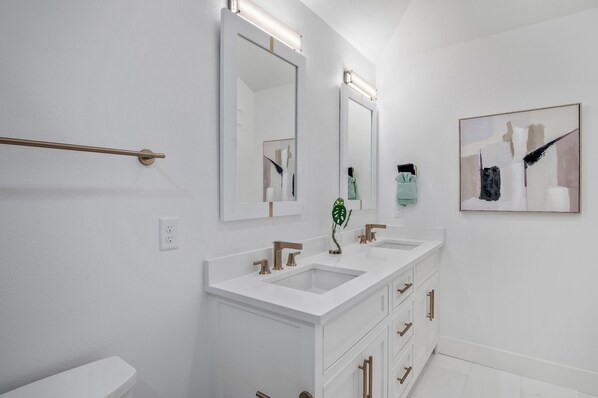 Un baño moderno con tocador blanco, dos lavamanos, un espejo grande y un cuadro abstracto enmarco.