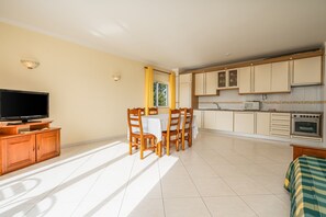 Un'ampia zona giorno con TV, tavolo da pranzo e sedie, una cucina con pensili ed elettrodomestici, e un letto con coperta a righe.