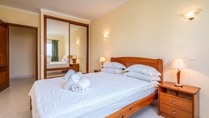 Ein ordentlich bezogenes Bett mit Kissen und einem gerollten Handtuch, ein Nachttisch aus Holz mit Lampe, Spiegel und Kleiderschrank.