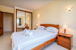 A neatly made bed with pillows and a rolled towel, a wooden nightstand with a lamp, a mirror, and a wardrobe. - Apartment 'Marachique 205' with Sea View, Shared Pool and Shared Garden (Alvor)