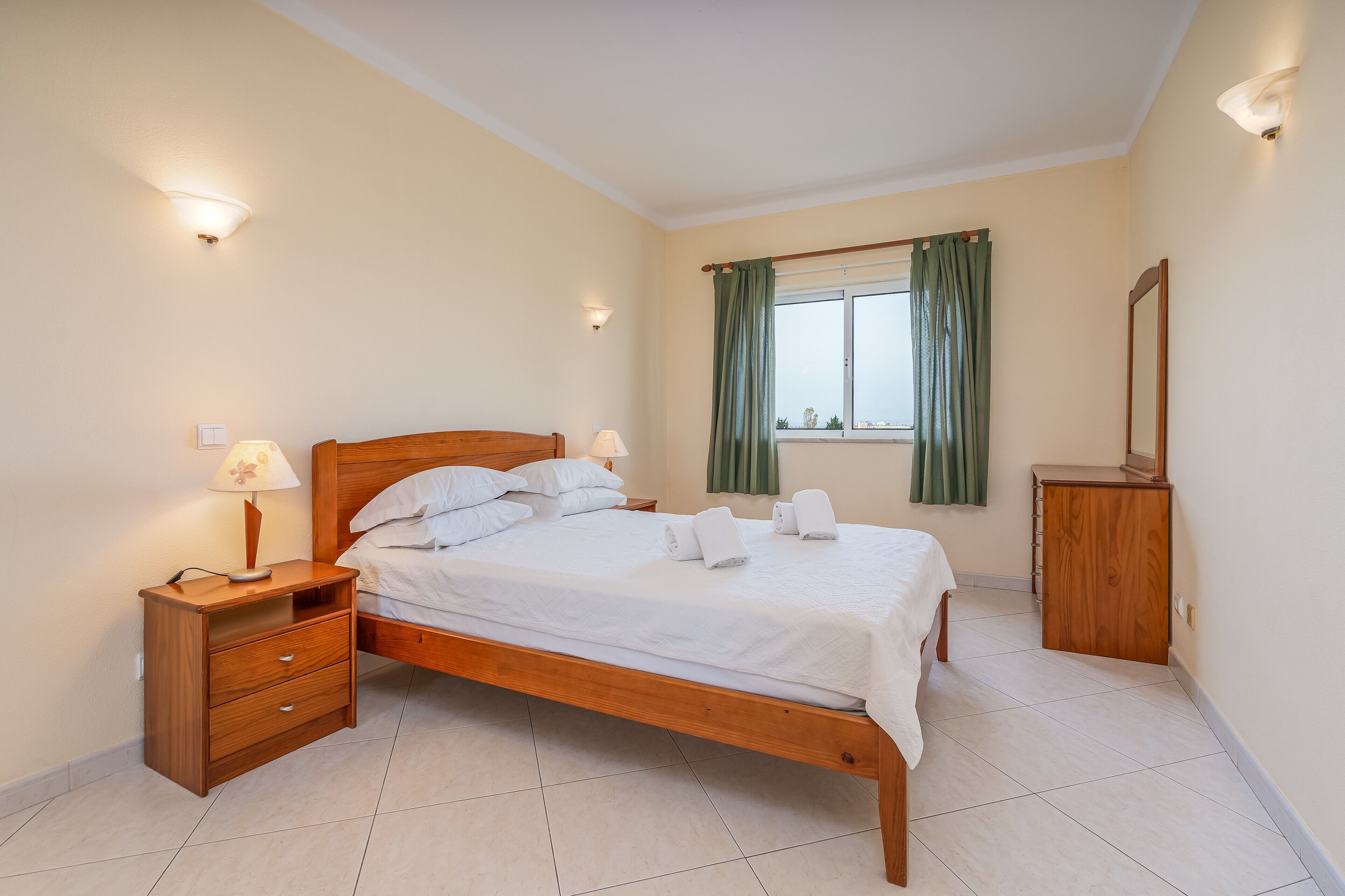 A neatly made bed with white linens, a wooden nightstand with a lamp, a dresser, and a window with curtains.