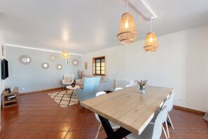 Un vaste espace de vie comprenant une table à manger en bois, des chaises blanches, un fauteuil bleu et un tapis à motifs.