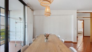 Un comedor con mesa de madera, sillas blancas, un jarrón con flores y una lámpara colgante.
