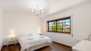 Un dormitorio con piso de madera, una cama con ropa de cama blanca, una araña, una mesita de noche con lámpara y una ventana con vista a follaje.