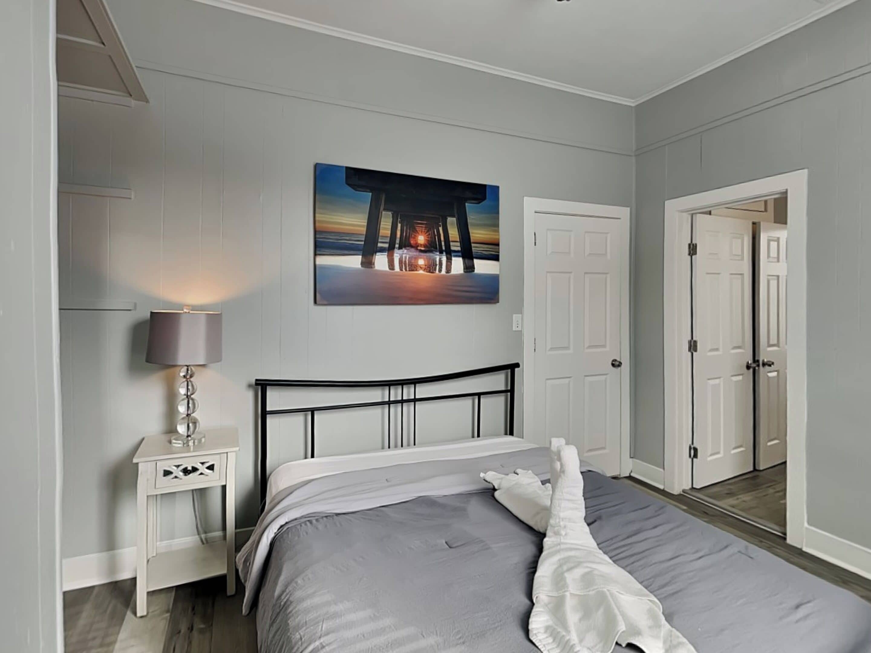 A bedroom with a bed, a nightstand with a lamp, a door, and a painting on the wall.