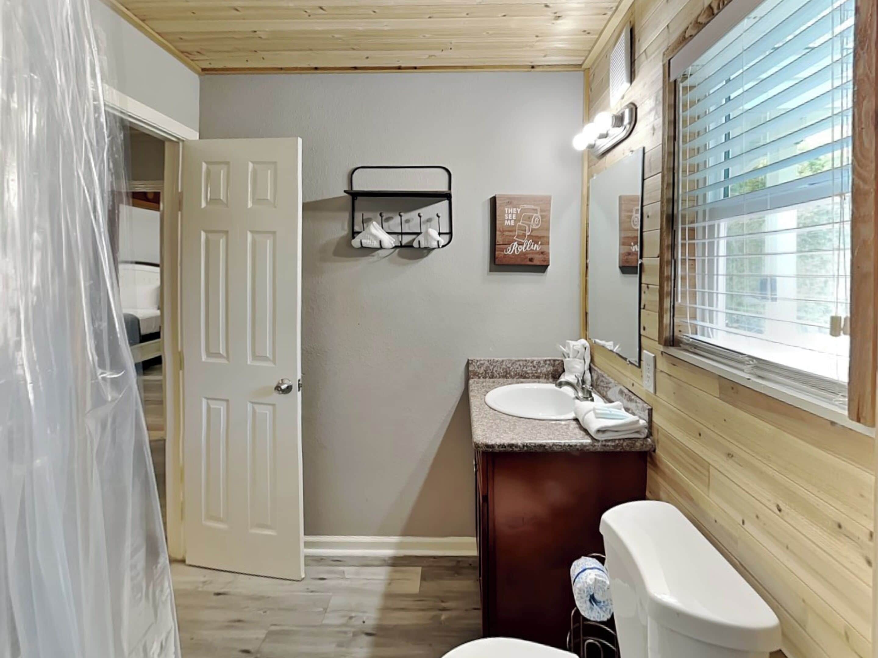 A bathroom with a wooden ceiling, a white door, a granite countertop sink, a mirror, and a window with blinds.