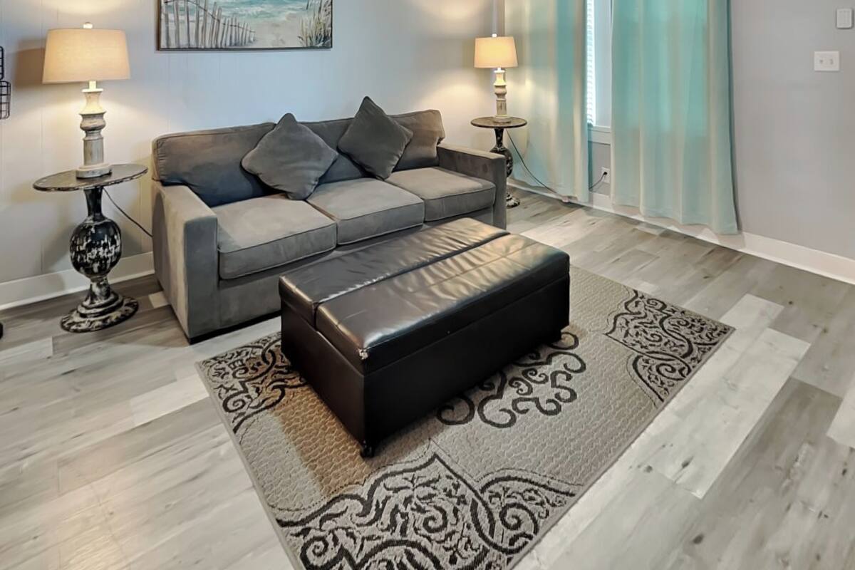 Una sala de estar con un sofá gris, dos mesitas de noche con lámparas, un reposapiés negro y una alfombra estampada.