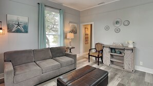 A living room with a grey sofa, a black ottoman, a wooden chair, and a small table with a lamp. - Pleasure Pier Retreat Manor by Ryson Vacation Rentals (Galveston)