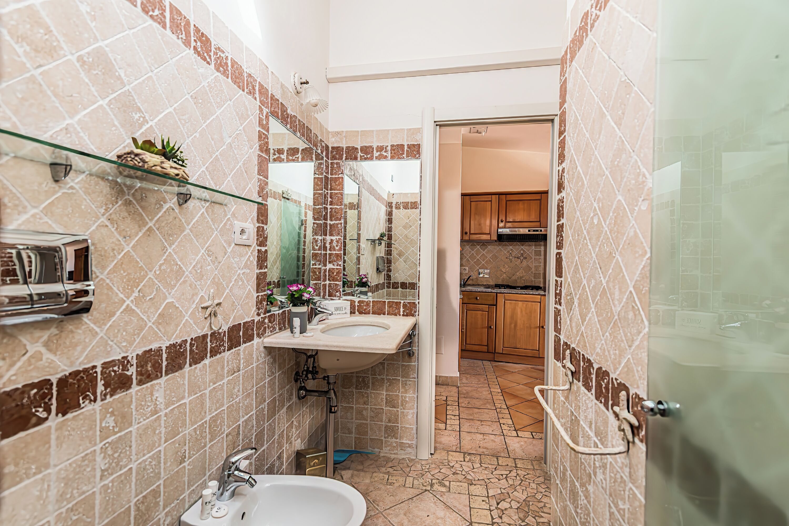 A bathroom with a sink, mirror, and shower. There is a small shelf with a plant and a toothbrush holder. The floor has a patterned tile design.