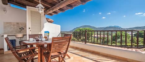 A balcony with a wooden table and chairs, a hanging light fixture, and a view of green hills.