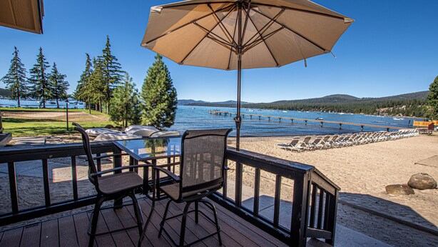 Une terrasse en bois avec une table et des chaises, un parasol, et une vue sur un lac où l’on peut apercevoir des bateaux et un quai.