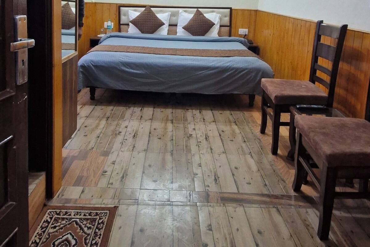 A bedroom with a wooden floor, a bed with a headboard, two wooden chairs, and a small rug.