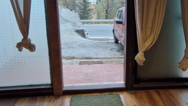 A room with a wooden floor, a green mat, and a view of a red car and buildings through a glass door with curtains. - White Villa (Shimla)
