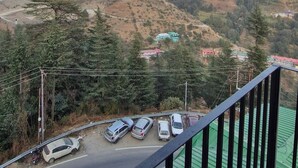 A balcony with a view of a hillside town, a road with parked cars, and a green-roofed building. - White Villa (Shimla)