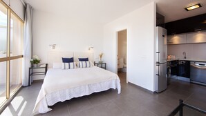 A modern hotel room with a large bed, a small bedside table with a lamp, a view through a large window, and a kitchenette area with a refrigerator and oven. - Studio in Marbella Steps From Sandy Beach (Marbella)