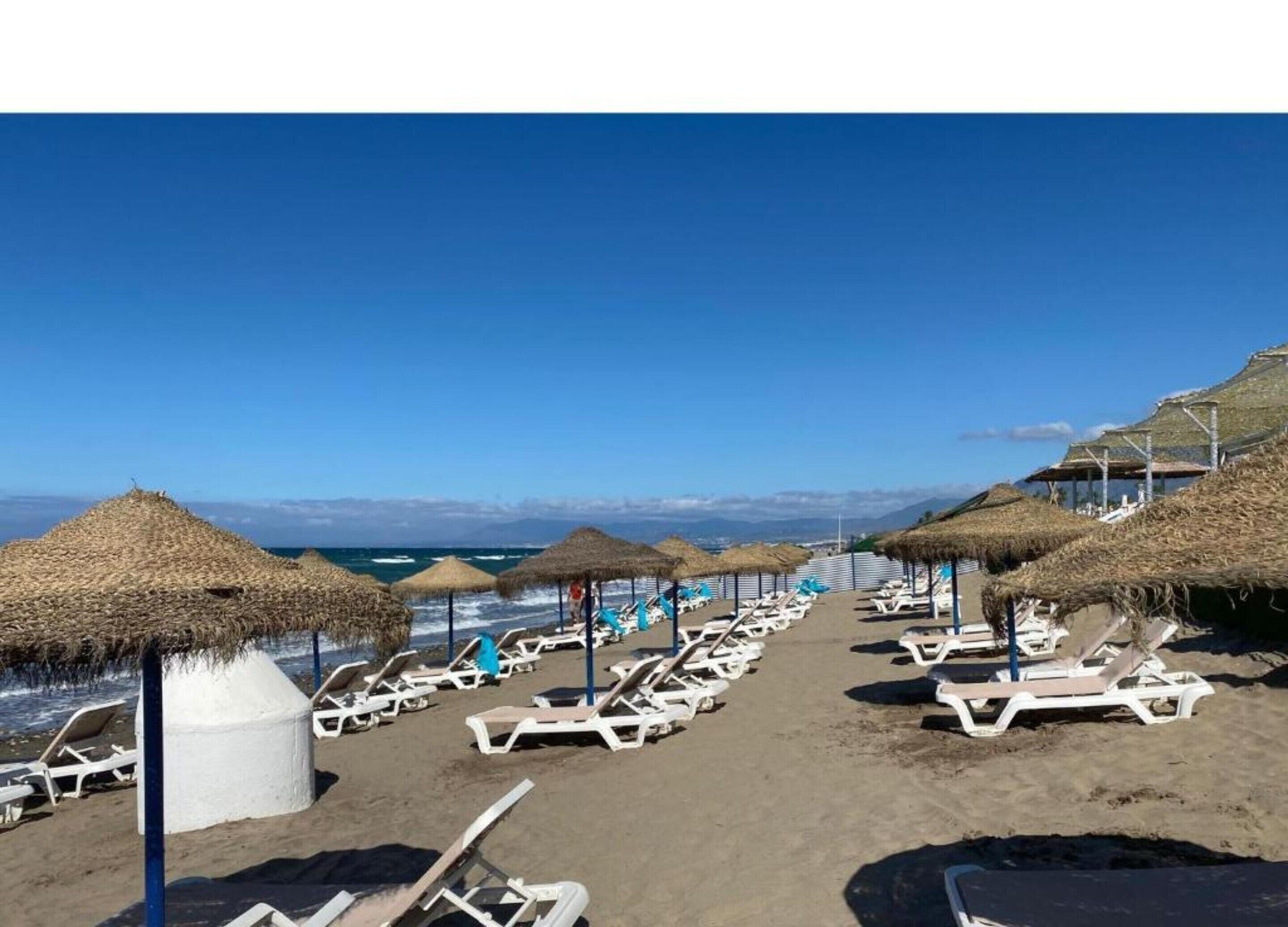 Ein Strand mit Reihen von Liegestühlen und Reetdachschirmen, einem klaren Himmel und Blick auf das Meer sowie ferne Berge.