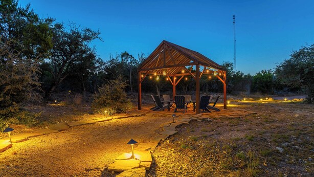 Um gazebo de madeira com cadeiras e luzes de corda, cercado por um caminho de cascalho e árvores.