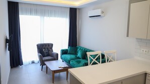 Uma sala de estar moderna com um sofá cinzento, um sofá verde, uma mesa de jantar branca e uma cadeira branca. Há uma janela com cortinas, um ar condicionado na parede e uma área de cozinha com armários brancos.