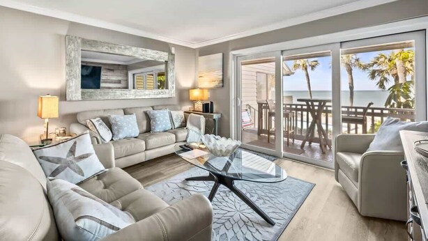 A modern living room with a sofa, coffee table, and a large window with a view of palm trees. - Aldea 18 Beachfront Condo in Belleair Beach (Belleair Beach)
