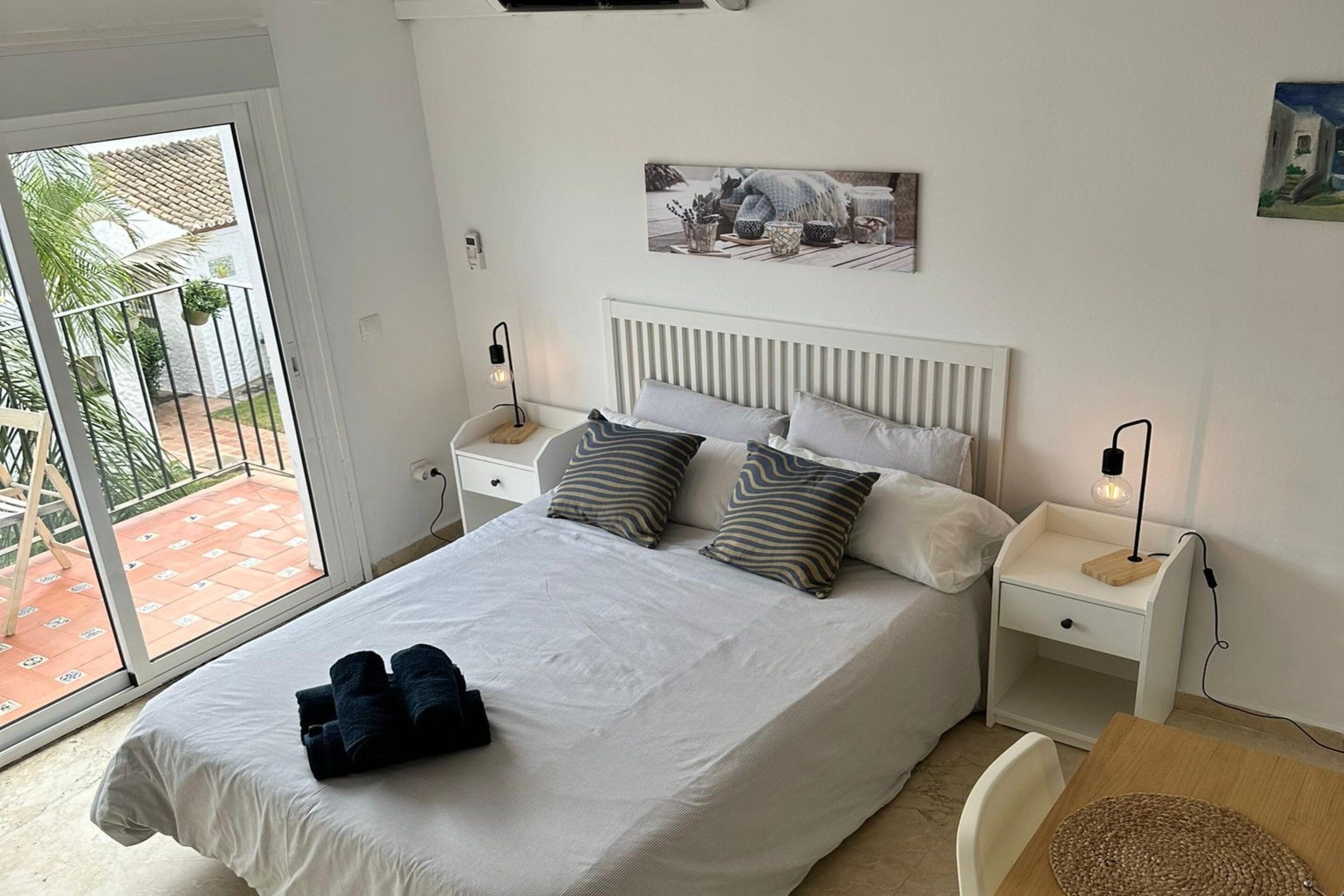 Un dormitorio ordenado con cama, almohadas, mesita de noche, lámpara y vistas al exterior.