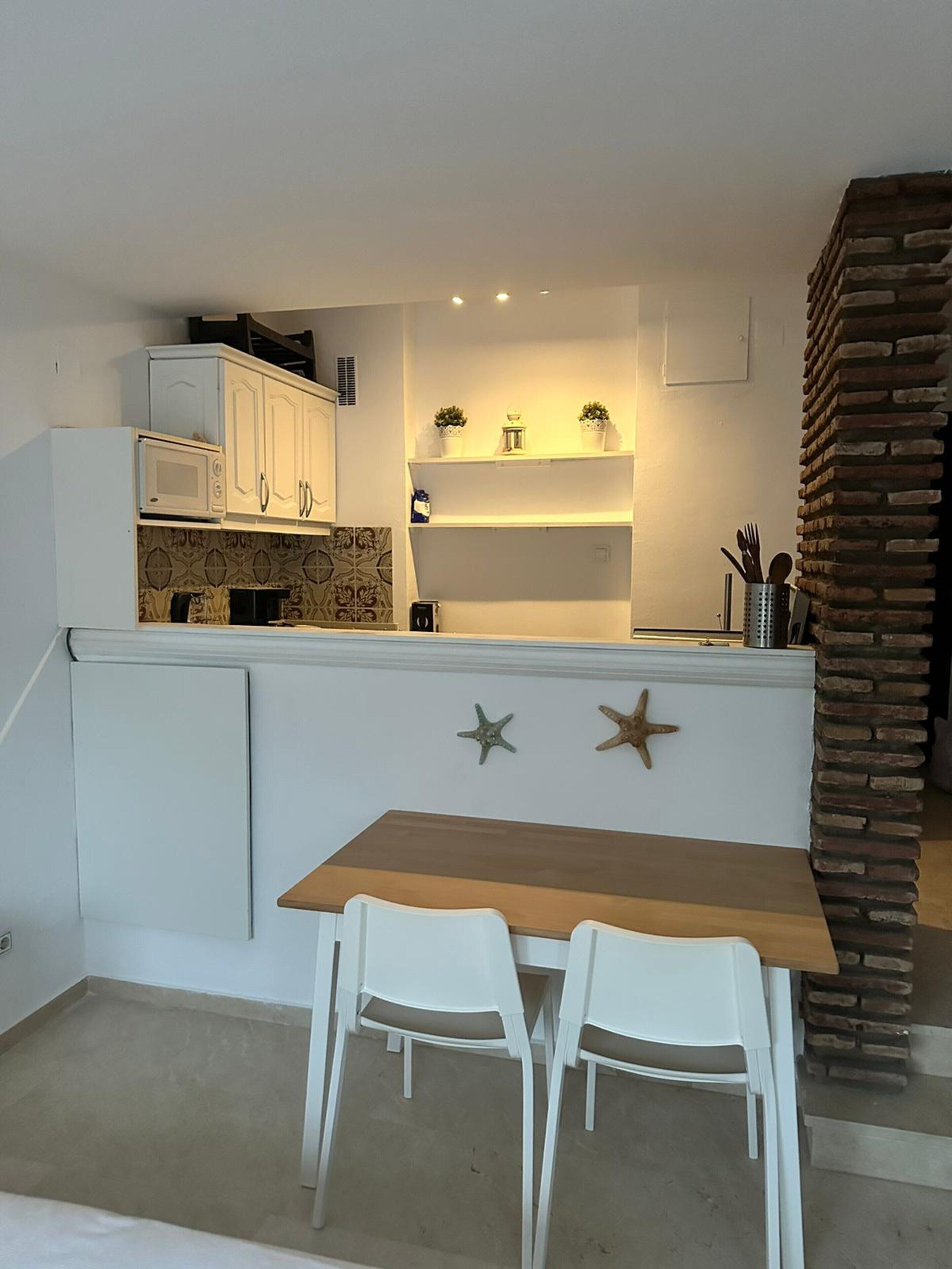 A compact kitchen with white cabinets, a wooden table, and two white chairs. There are decorative starfish on the wall and a brick accent wall.