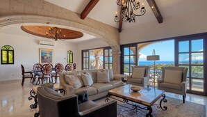 Un salon spacieux avec un grand canapé, des fauteuils et une table basse. Il comprend également un coin repas avec une table et des chaises, ainsi que de grandes fenêtres offrant une vue sur l’océan.