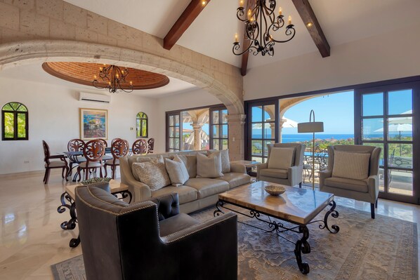 A spacious living room with a large sofa, armchairs, and a coffee table. There is a dining area with a table and chairs, and large windows offering a view of the ocean.