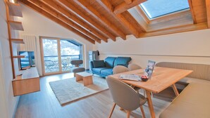 A modern loft-style room with a wooden ceiling, a large window, a sofa, a dining table, and a TV. - Apartment Jua (Zermatt)