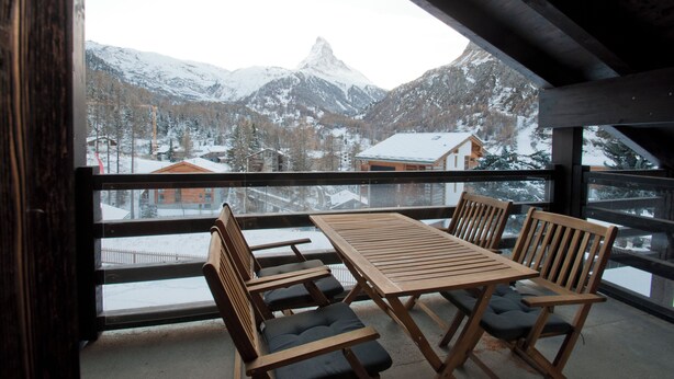 A wooden table and chairs on a balcony with a snow-covered mountain view. - Apartment Jua (Zermatt)