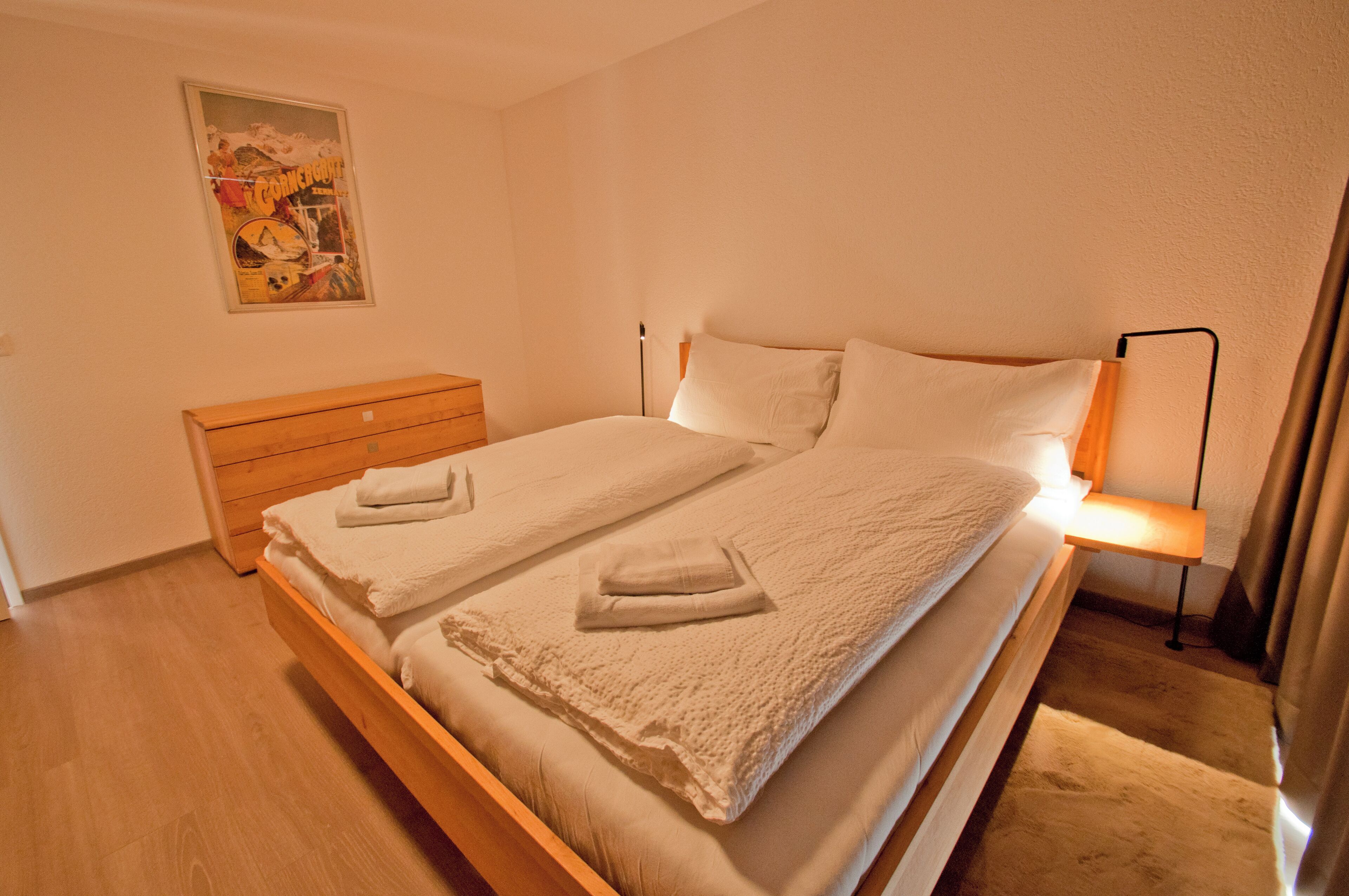 A double bed with white linens, a wooden nightstand, a wall-mounted lamp, and a poster on the wall.