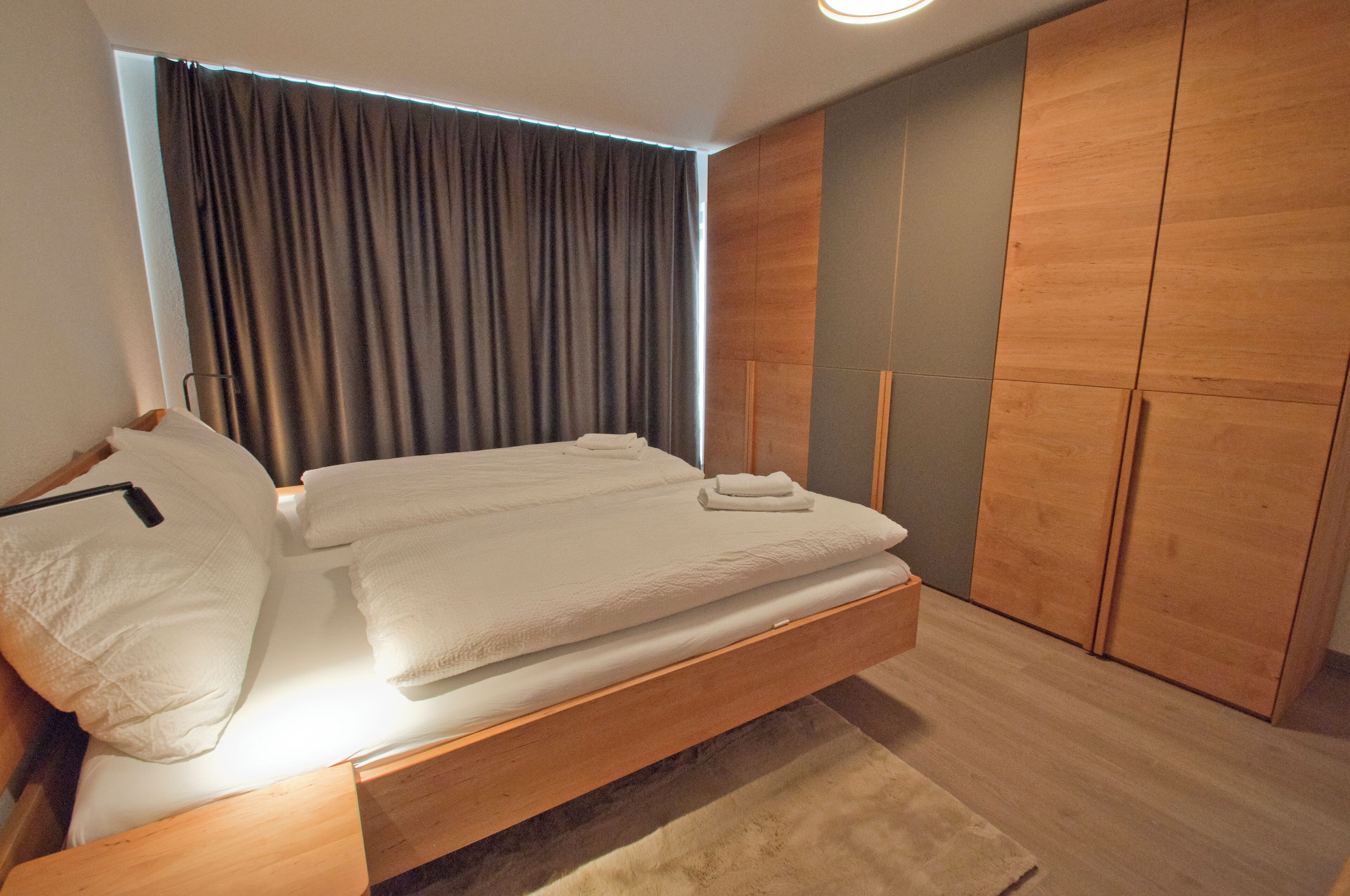 Un dormitorio moderno con una cama de madera, un armario grande y vistas de la ciudad a través de una ventana con cortinas.