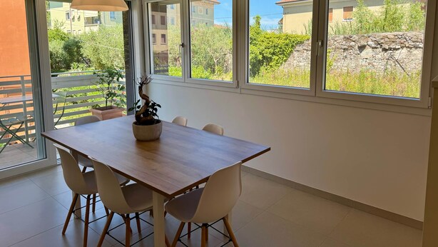 A dining area with a wooden table and chairs, a potted plant, and large windows with a view of buildings and greenery. - Domus Aurus Family, Domus Aurus Family  (Bardolino)