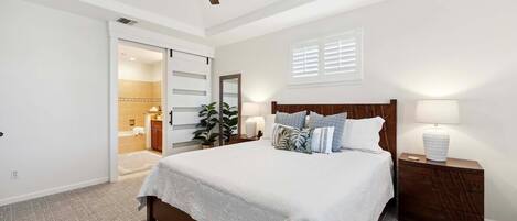 A bedroom with a large bed, a wooden headboard, a nightstand with a lamp, a ceiling fan, and a bathroom visible through an open door.
