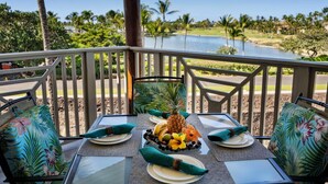 A dining table set for four with tropical-themed cushions, a fruit bowl, and a view of a golf course and water. - Waikoloa Beach Villas F32 (Waikoloa)