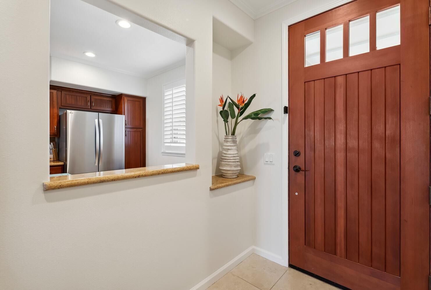 A hallway with a wooden door, a mirror reflecting a kitchen, and a plant on a shelf.