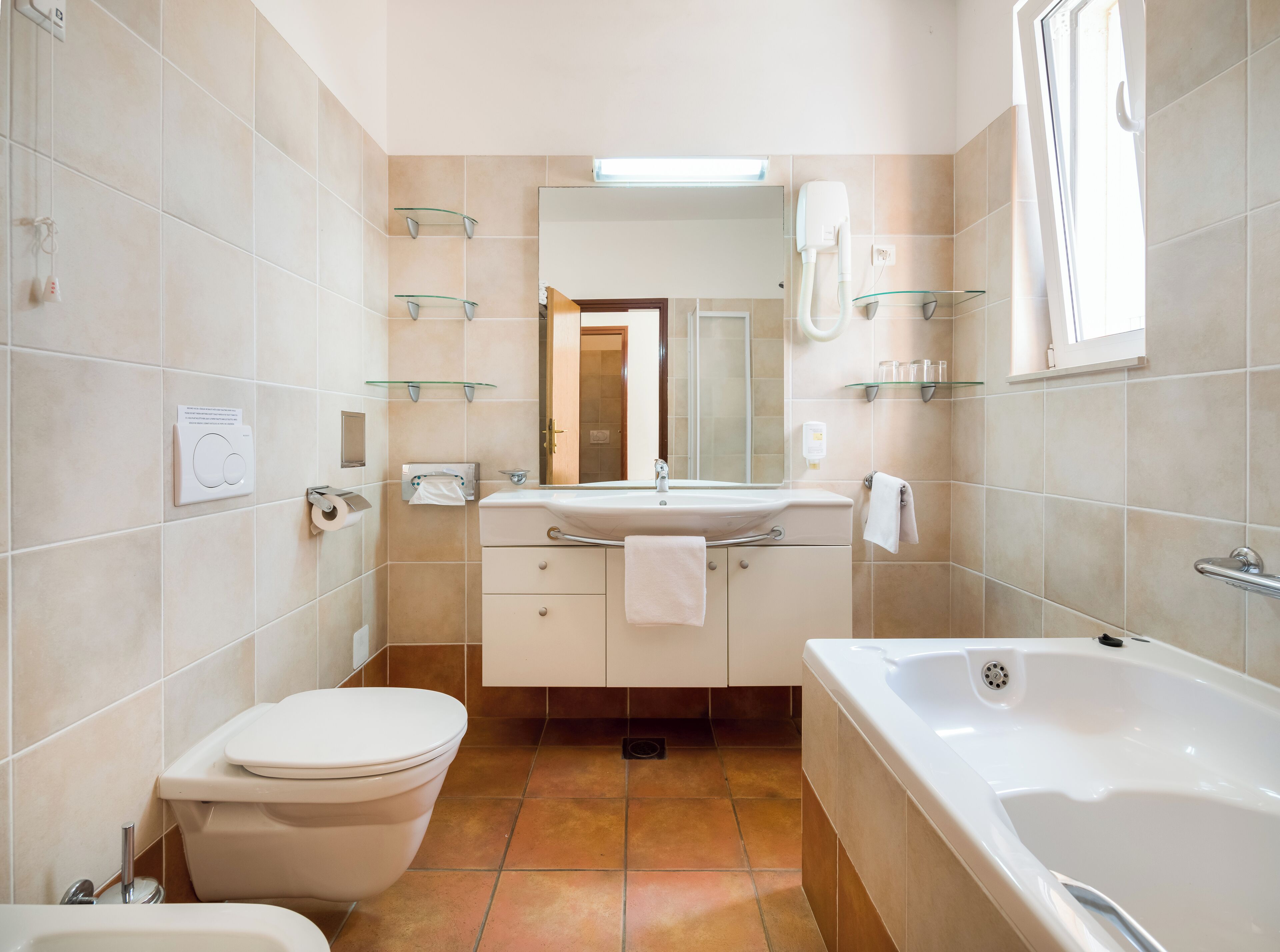 A bathroom with a toilet, sink, and bathtub. There are glass shelves above the sink and a mirror above the bathtub.