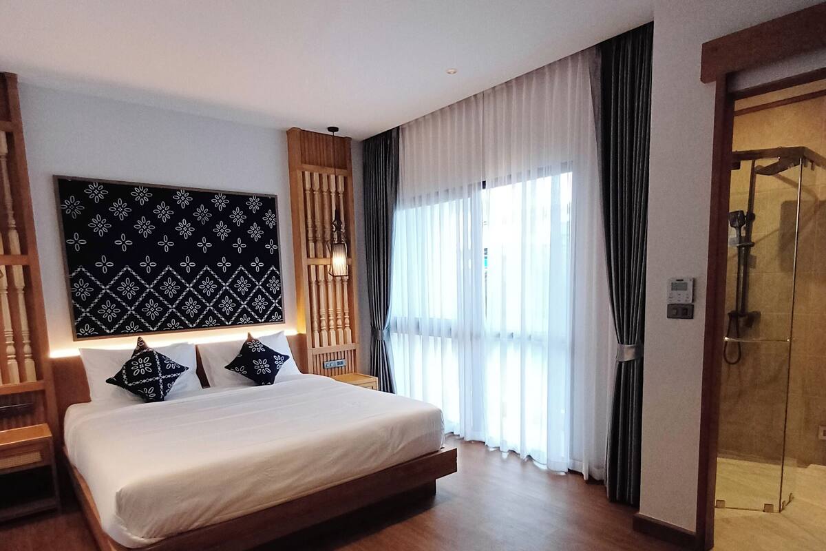 Ein modernes Hotelzimmer mit einem großen Bett, einem Holz-Kopfteil, einer Wandleuchte sowie einer Dusche und einem gemusterten Wandteppich.