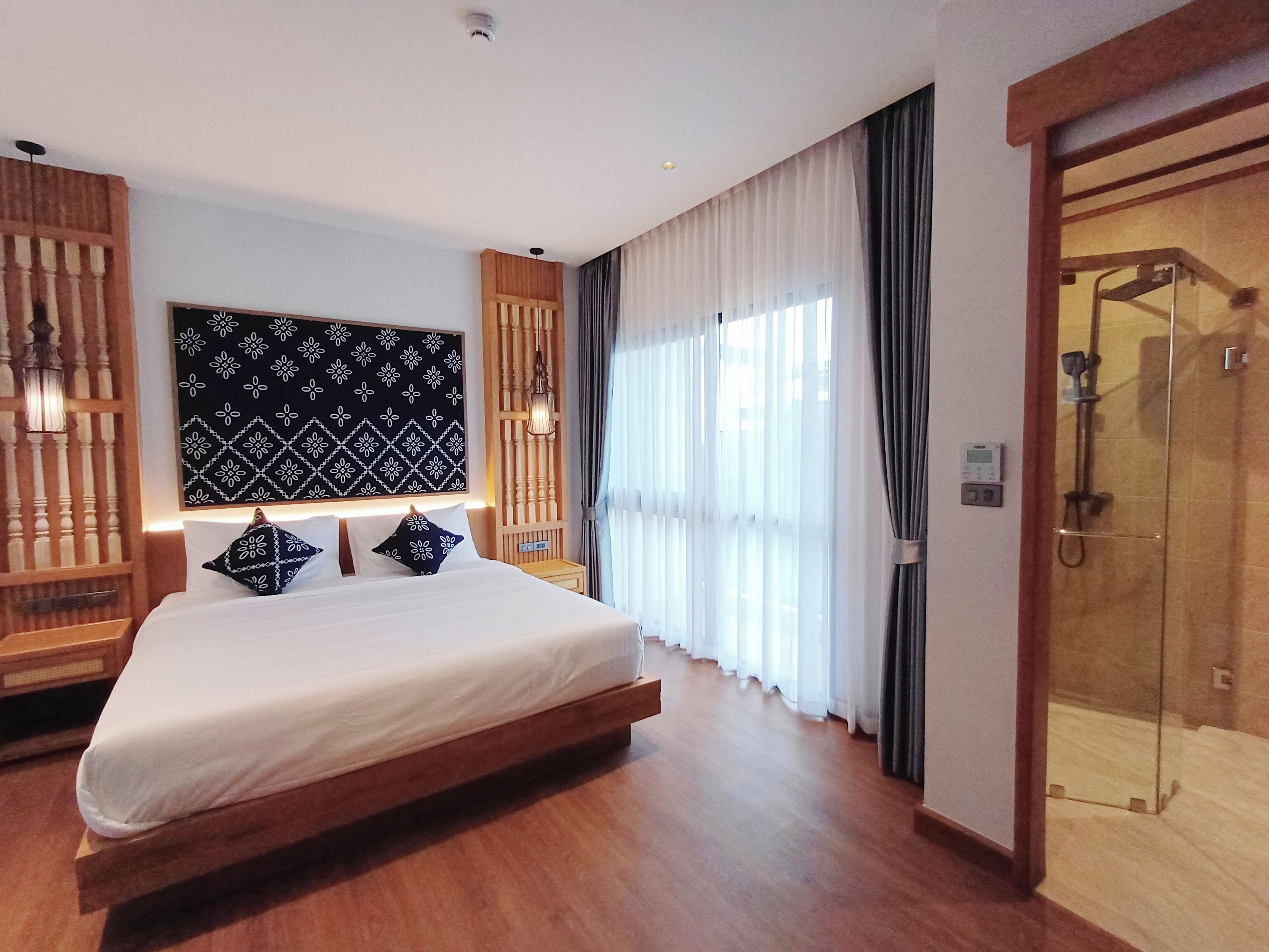 A modern hotel room with a large bed, a wooden headboard, a large window with sheer curtains, and a glass-enclosed shower area.