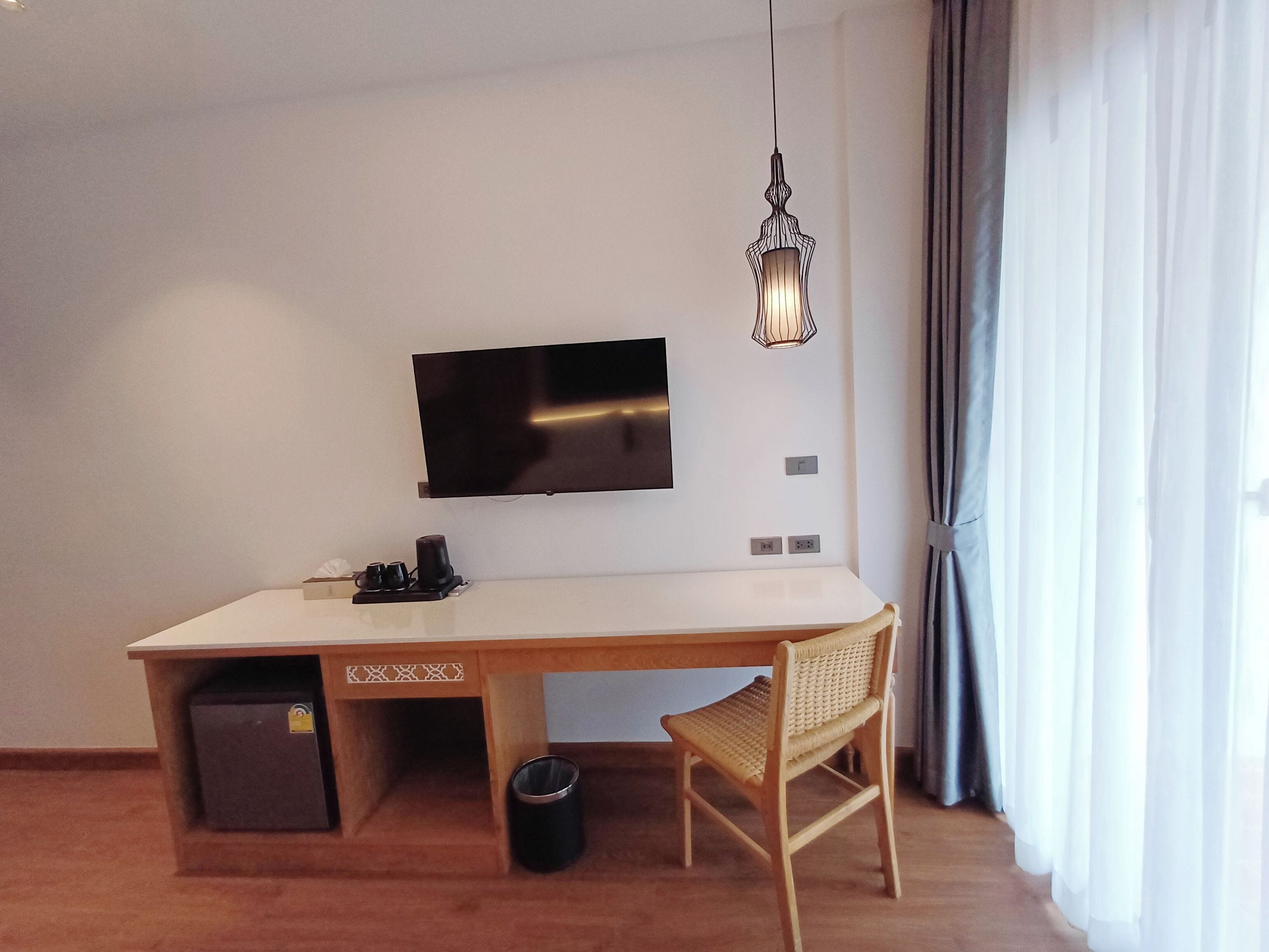 Ein modernes Zimmer mit Flachbildfernseher an der Wand, einem Holztisch mit Stuhl, einer kleinen Mülltonne und einer dekorativen Pendelleuchte.