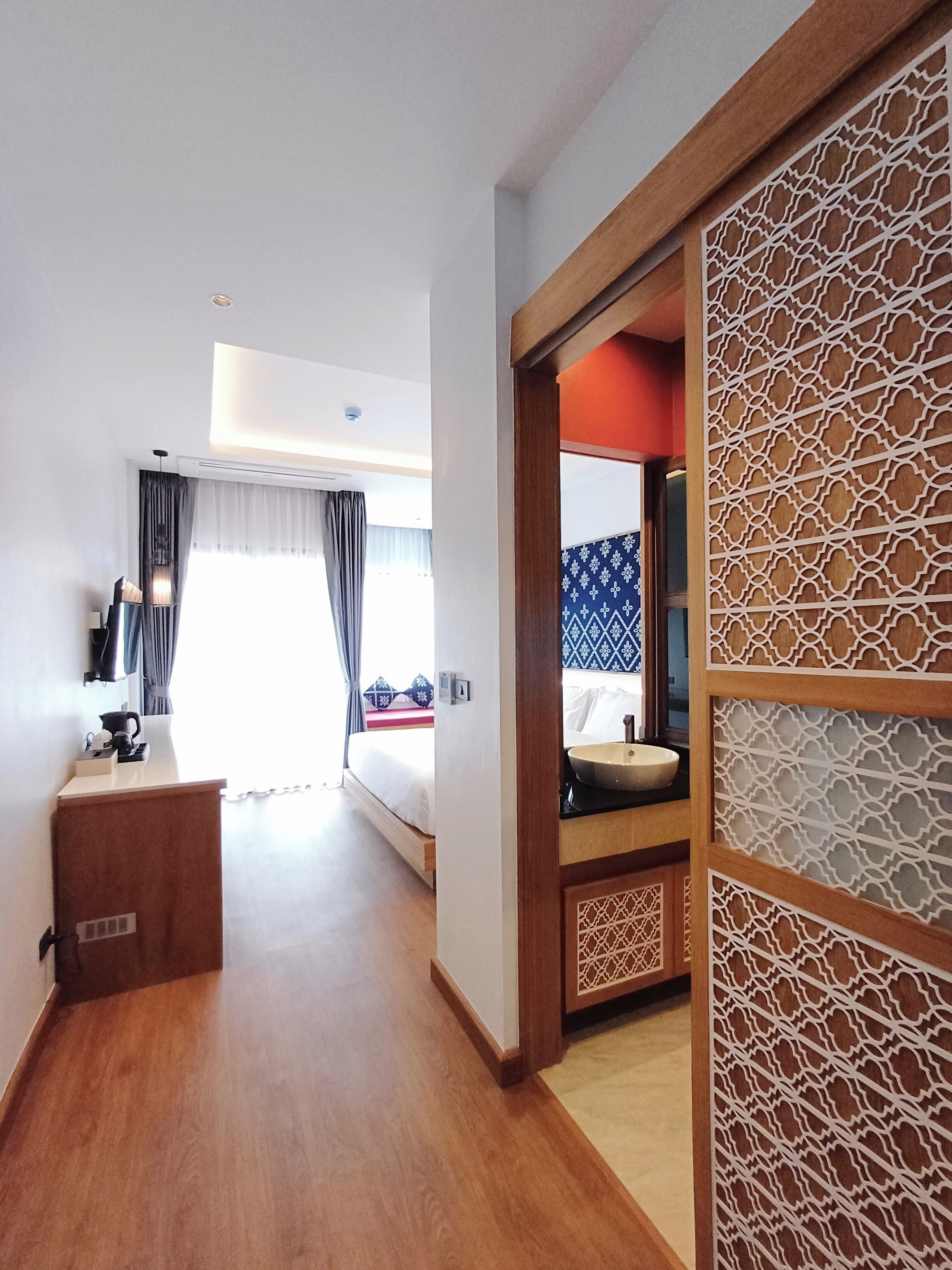 A hotel room with a wooden floor, a bed with a blue and white patterned headboard, a bedside table with a lamp, and a decorative screen with intricate patterns.