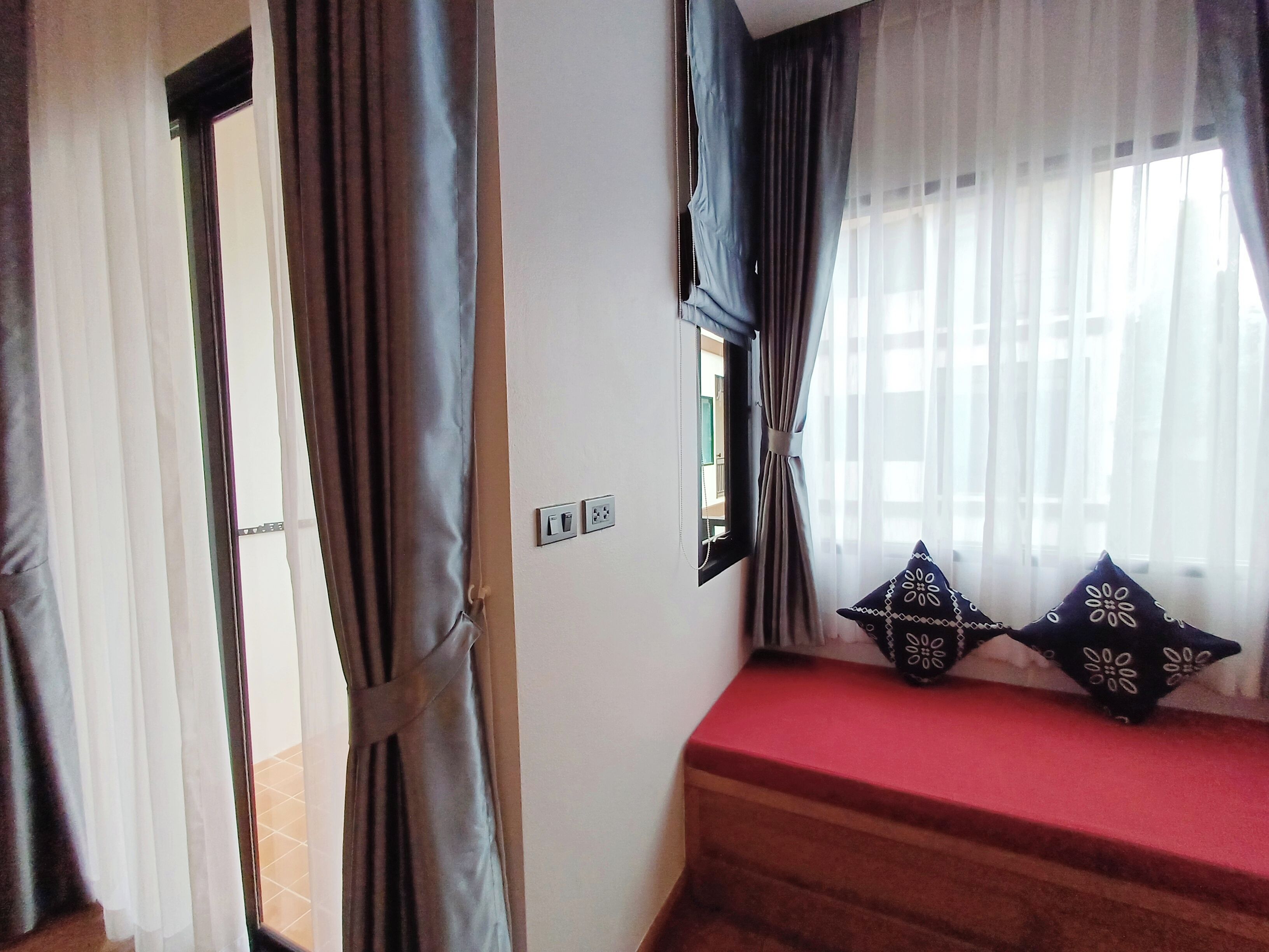 A room with a window, curtains, a red bench, and a wall socket.