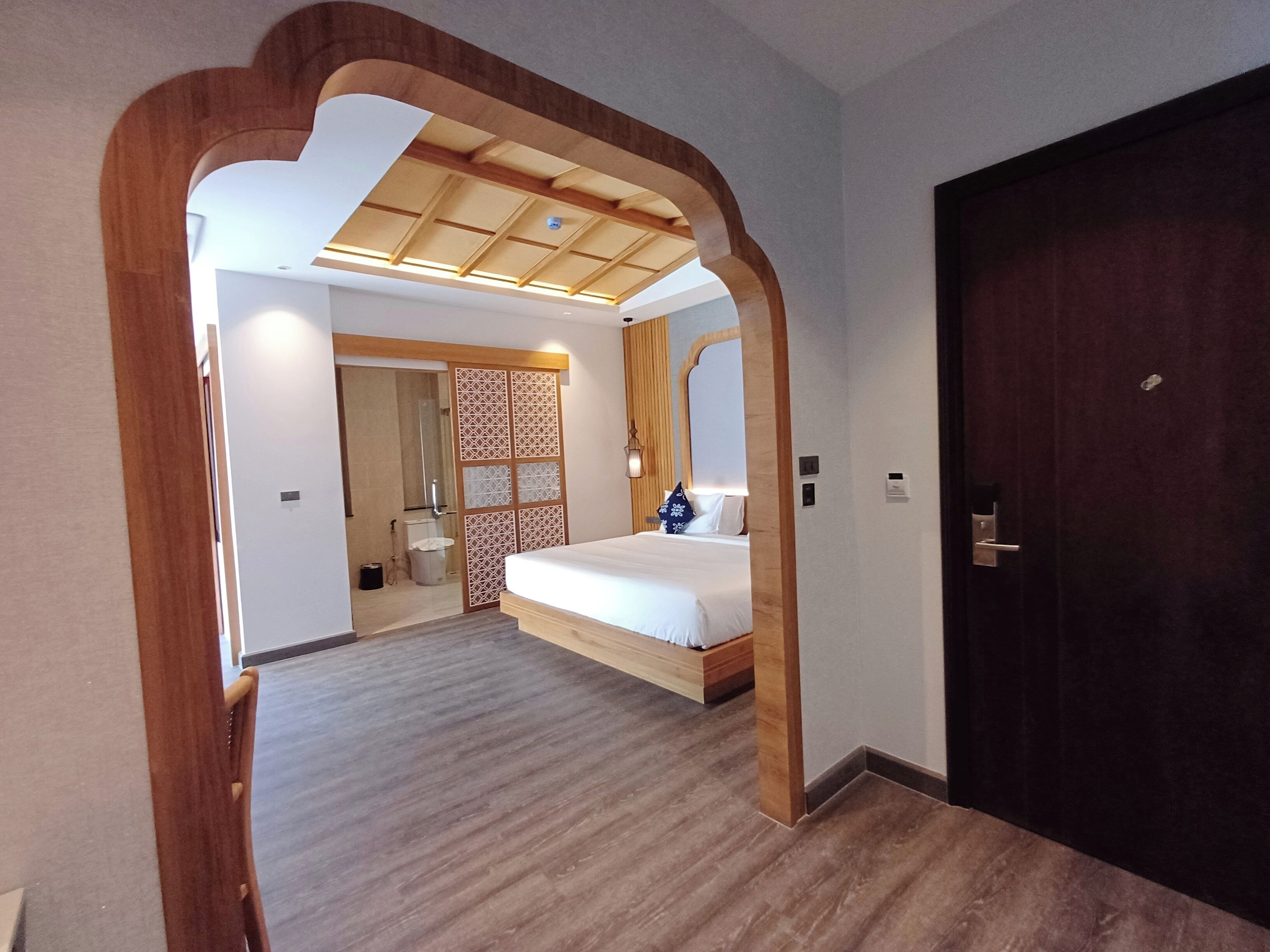 A hotel room with a wooden floor, a bed with white bedding, a wooden door, and a view of the bathroom.