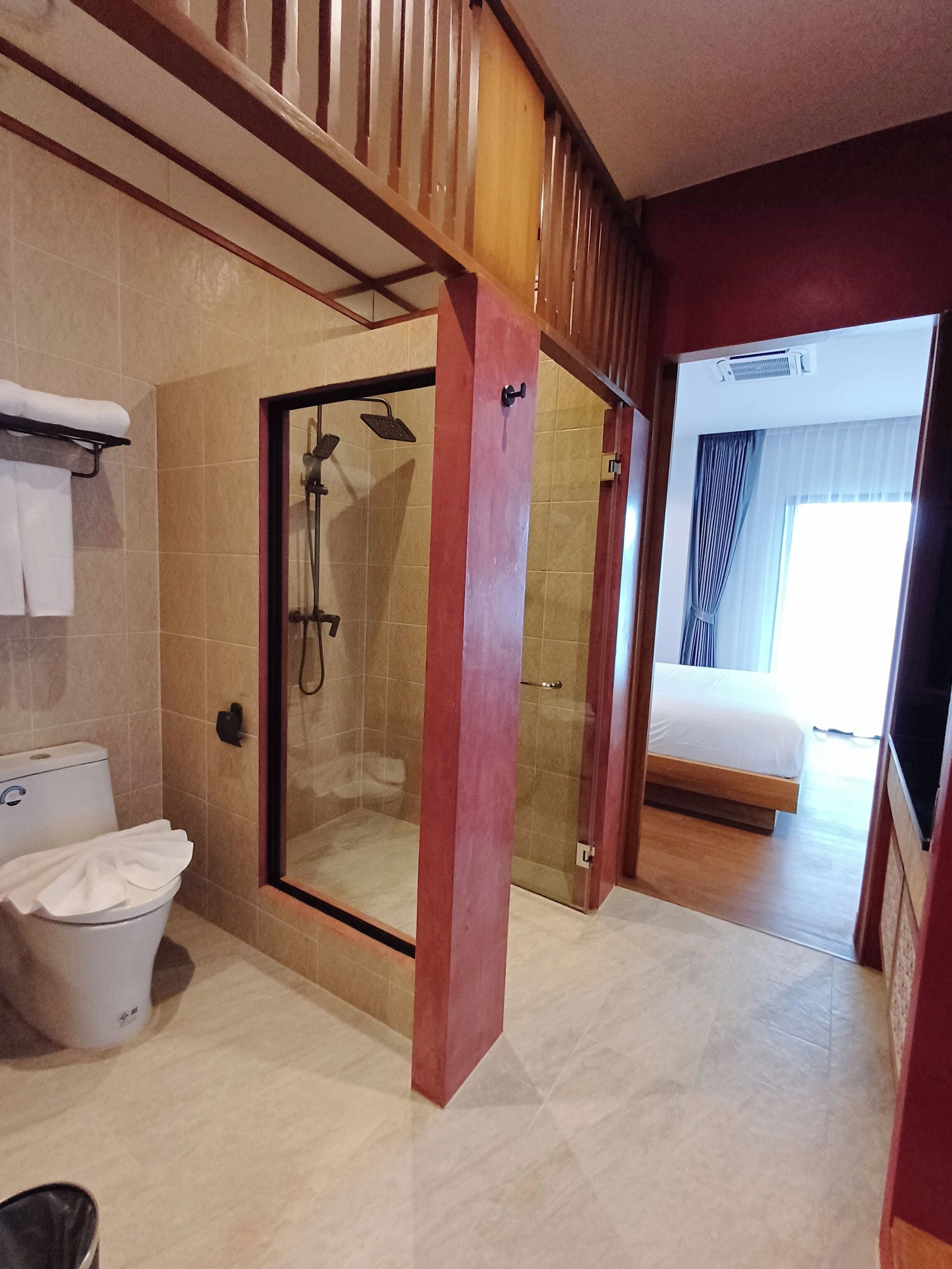 A hotel bathroom with a glass-enclosed shower, a toilet, and a bed visible through an open door.
