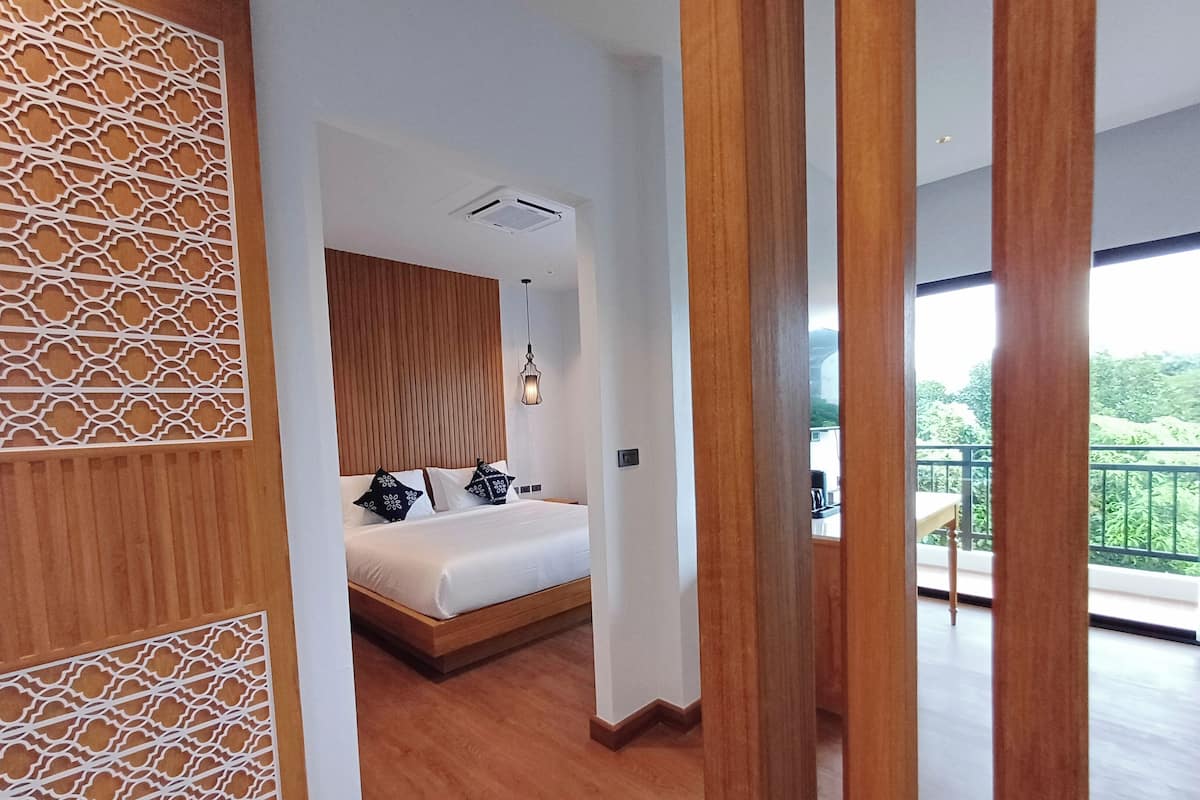 Ein modernes Schlafzimmer mit einem Holzkopfende, einem Bett mit weißer Bettwäsche und gemusterten Kissen, einer Pendelleuchte und Blick auf einen Balkon mit Gartenmöbeln.